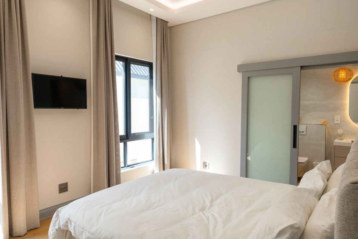 A modern bedroom featuring a sliding frosted glass door leading to an en-suite bathroom, large windows with curtains, and recessed ceiling lighting.