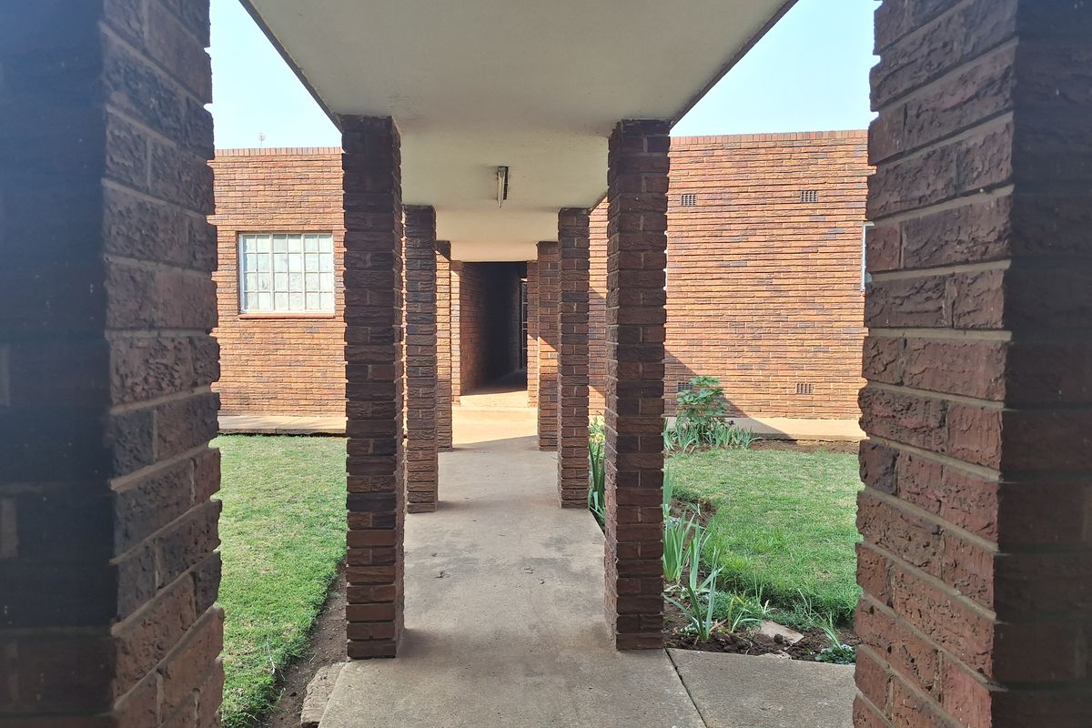 A covered concrete walkway framed by brick columns, leading through a residential courtyard with established lawn and brick building facades.