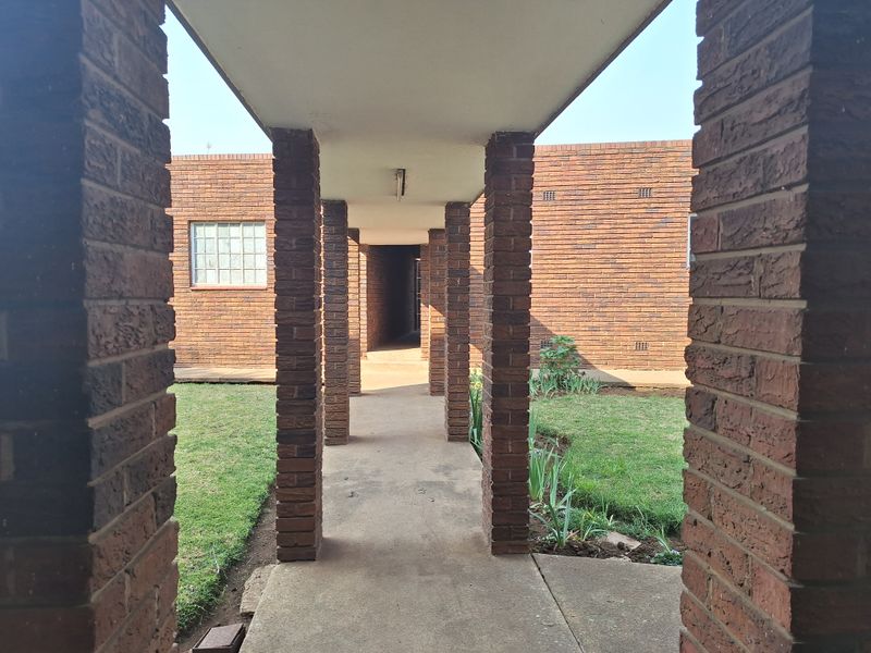 A covered concrete walkway framed by brick columns, leading through a residential courtyard with established lawn and brick building facades.