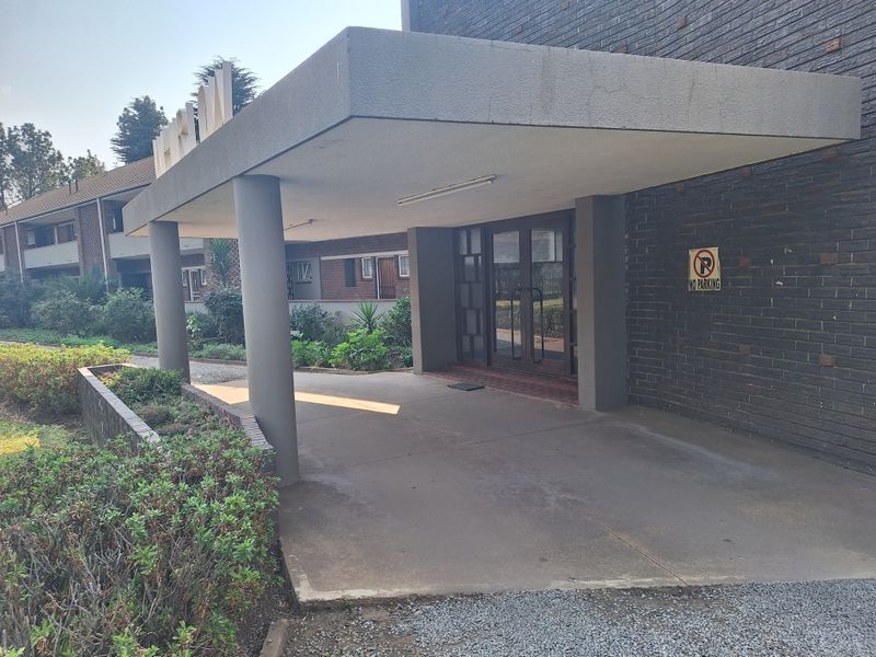 The apartment building features a covered entrance with concrete pillars, brick facade, and glass double doors, providing a sheltered approach to the main building.