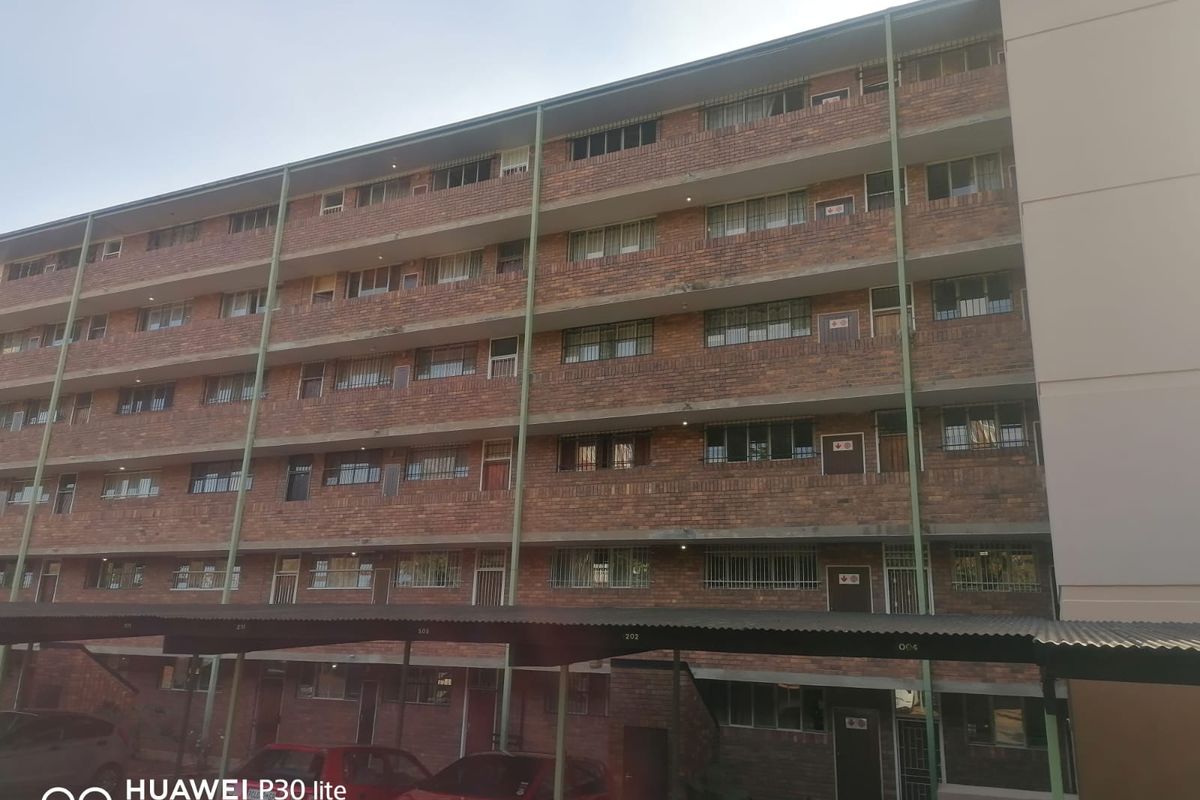 Exterior view of a multi-story brick apartment building featuring covered parking bays and security bars on windows.