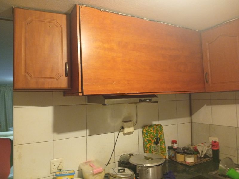 A dated kitchen area featuring wooden cabinetry, a tiled backsplash, and an integrated extractor fan, showing signs of wear and misaligned fixtures.