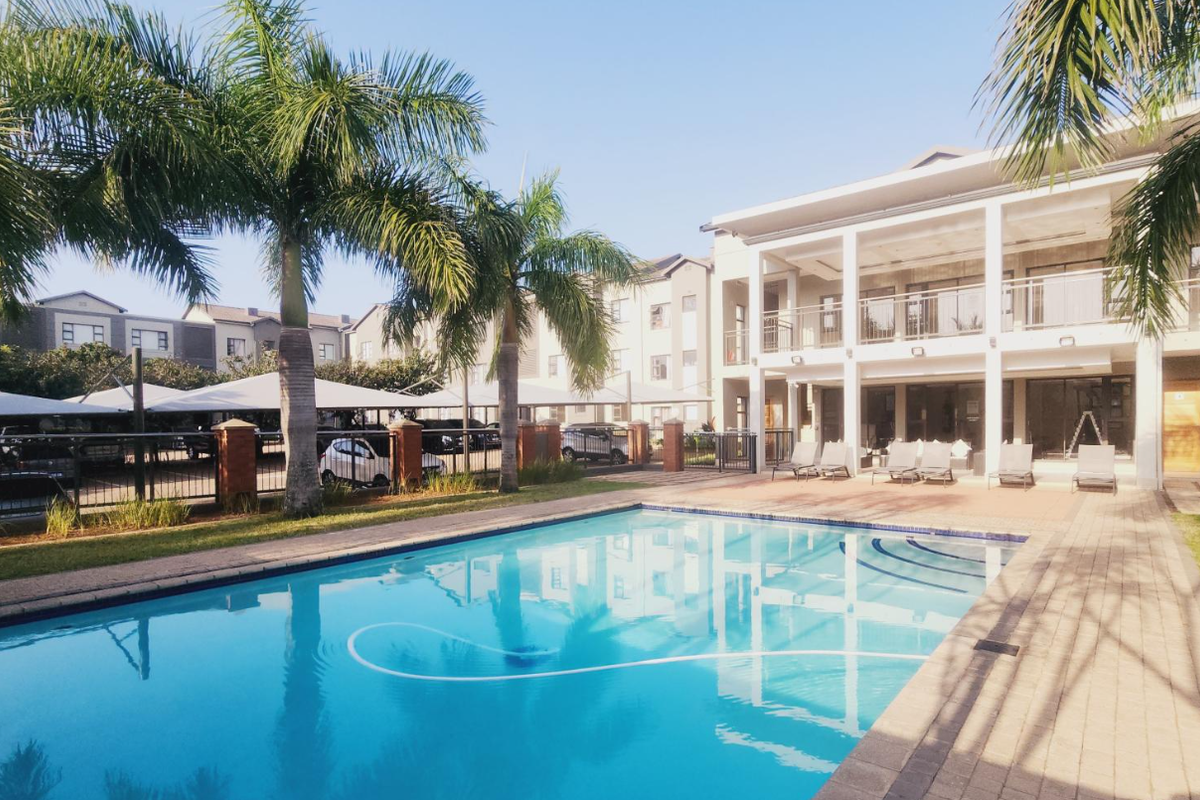 A well-maintained communal swimming pool area featuring paved decking, landscaped palm trees, and a modern multi-story apartment building in the background.