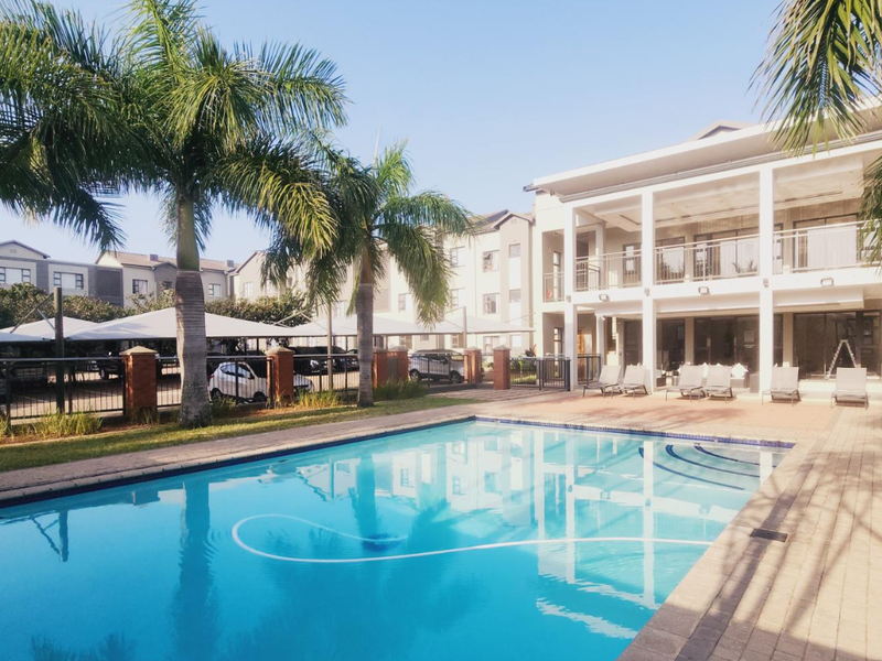 A well-maintained communal swimming pool area featuring paved decking, landscaped palm trees, and a modern multi-story apartment building in the background.