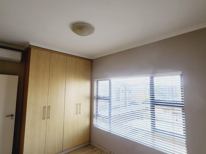 A bedroom featuring built-in wooden cupboards, a wall-mounted air conditioning unit, and a large window fitted with horizontal blinds.