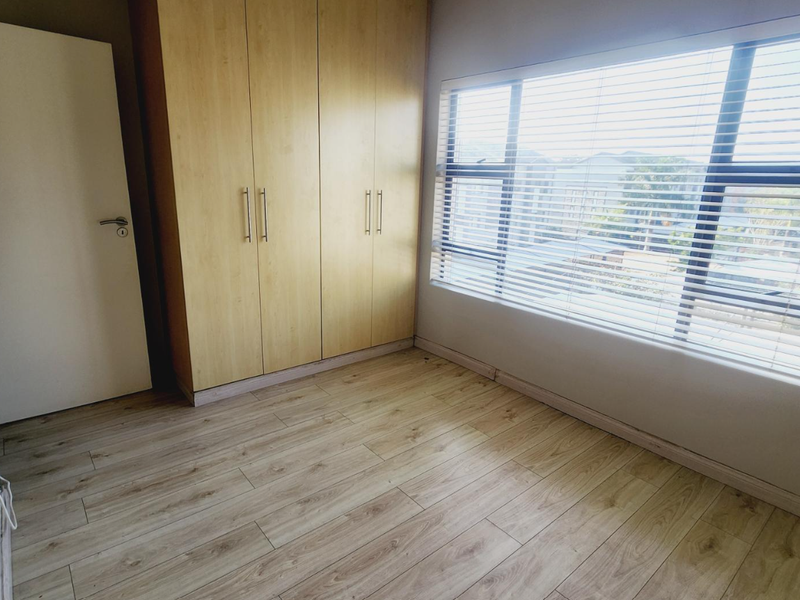 A bright bedroom featuring light wood-look laminate flooring, built-in wooden cabinetry, and large windows fitted with horizontal blinds.