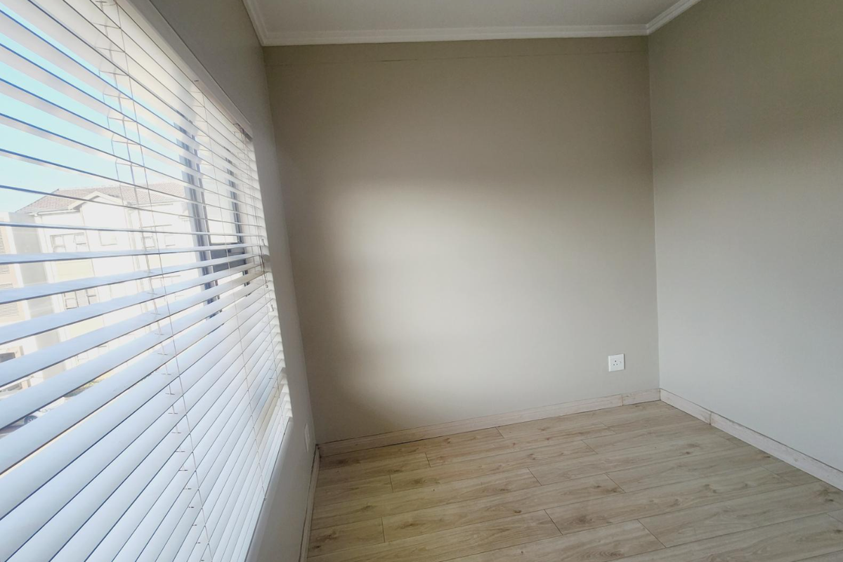 A bright, empty bedroom featuring light wood-look laminate flooring, neutral wall tones, and fitted venetian blinds.
