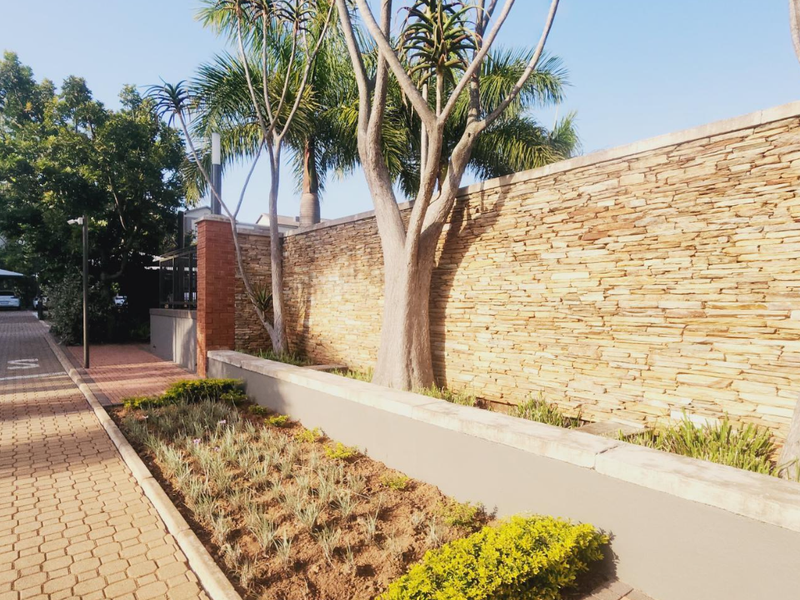 A well-maintained exterior view of the apartment complex featuring a stone-clad boundary wall, paved driveway, and landscaped garden beds with mature trees.