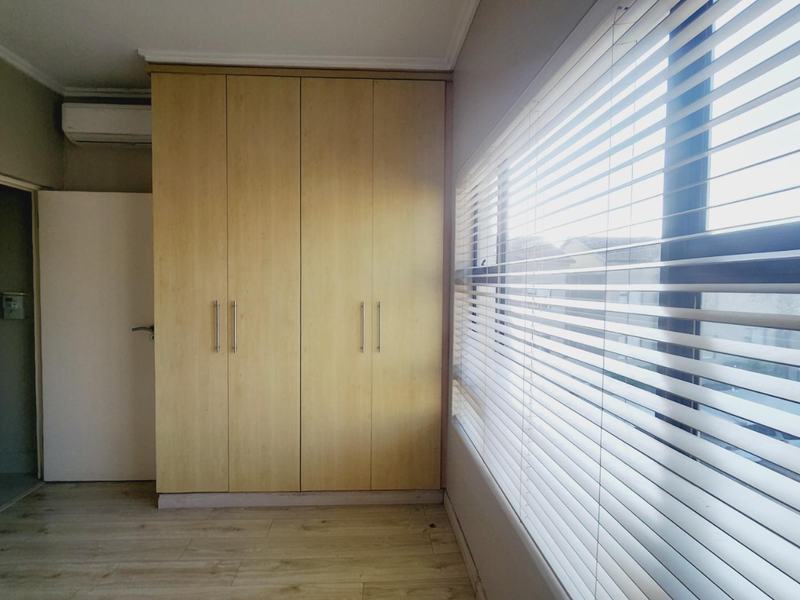 A bright bedroom featuring built-in wooden cabinetry, light-toned laminate flooring, and large windows fitted with horizontal blinds.
