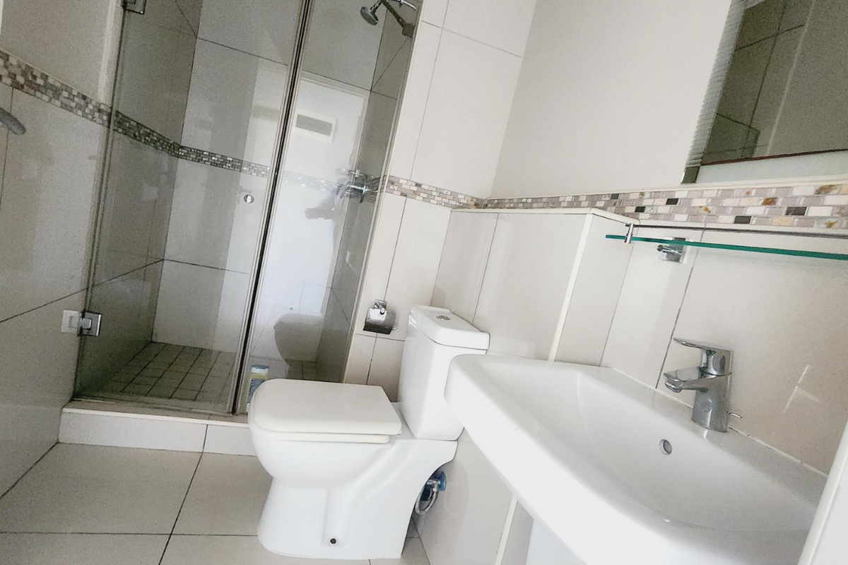 A functional bathroom featuring a glass-enclosed shower, a pedestal basin with chrome fixtures, and neutral wall tiling with a decorative mosaic accent border.