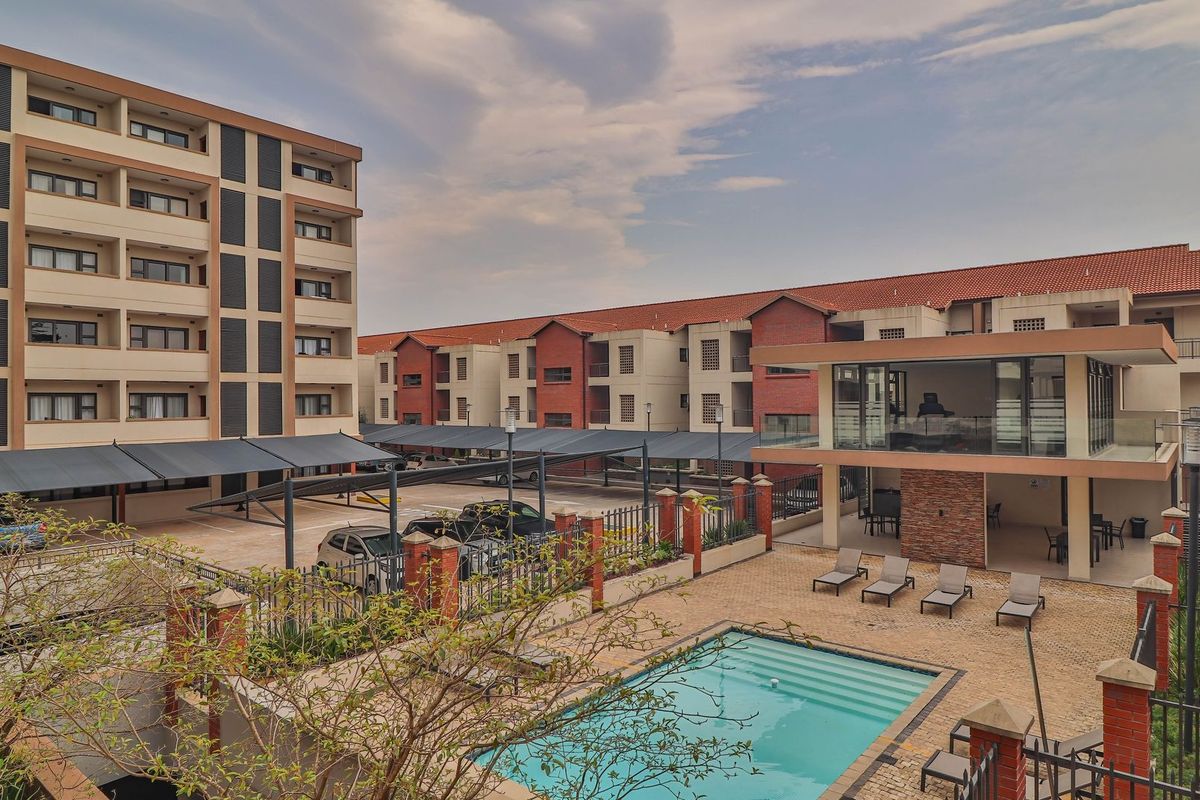 Elevated view of the apartment complex featuring a swimming pool, paved pool deck, covered parking bays, and a multi-story residential building.