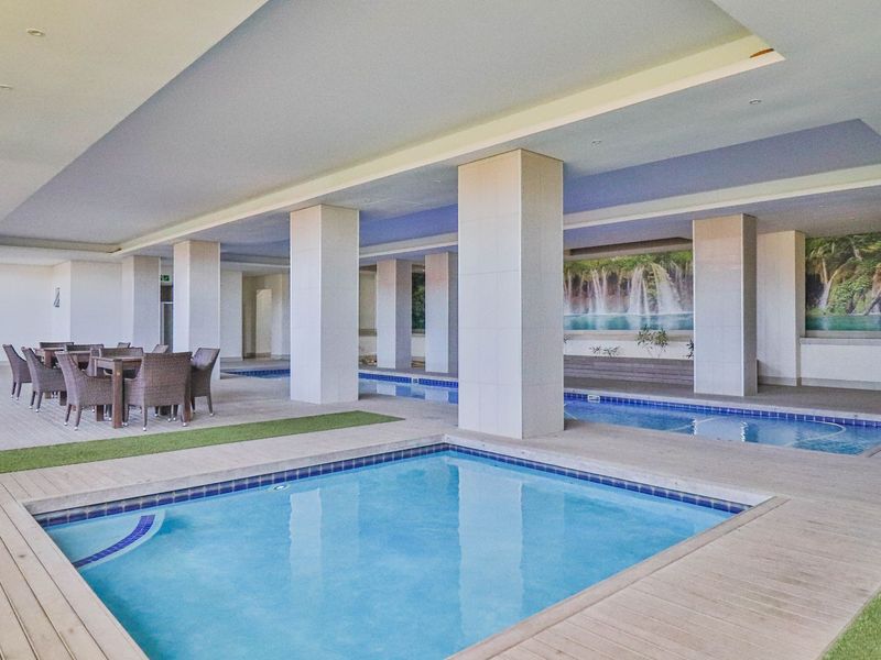 A spacious indoor pool area featuring multiple swimming pools, tiled decking, and structural columns, creating a modern communal leisure space.