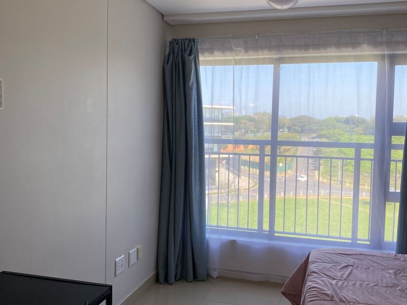 A bright bedroom featuring large windows with natural light, tiled flooring, and neutral wall finishes, offering a pleasant view of the surrounding area.