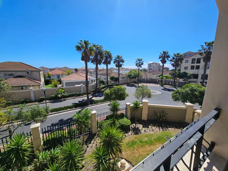 Elevated view overlooking the residential complex, featuring landscaped gardens, paved driveways, and surrounding apartment buildings under a clear blue sky.