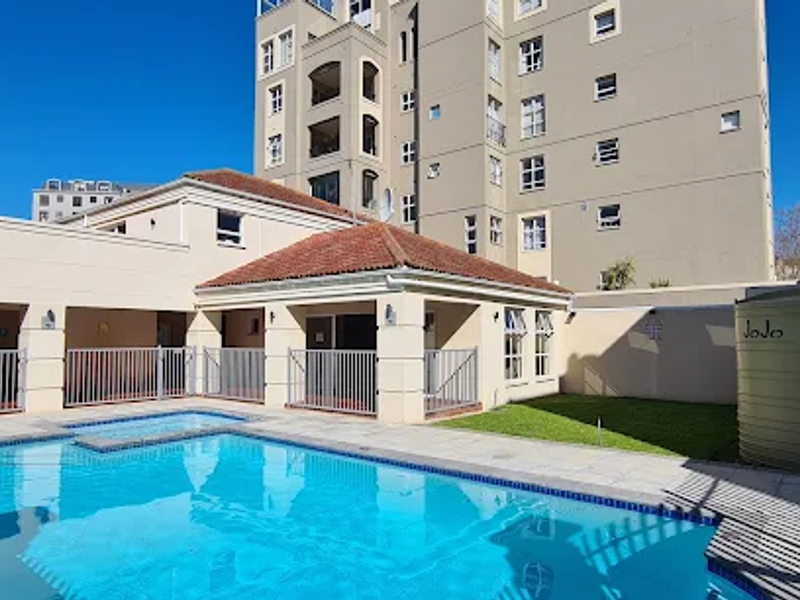 A well-maintained communal pool area featuring a paved deck, metal fencing, and a large water storage tank set against a multi-story apartment building.