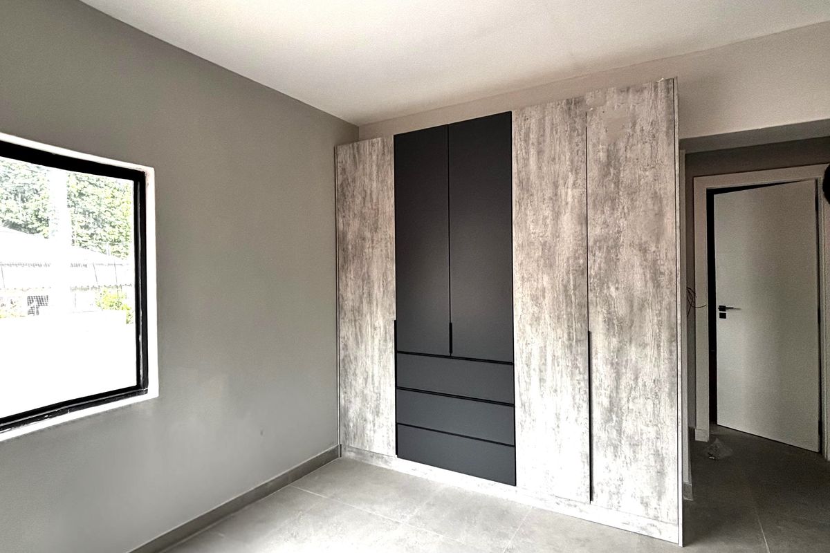 A modern bedroom featuring built-in floor-to-ceiling wardrobes with a mix of matte black and textured wood-look finishes, complemented by neutral tiled flooring.