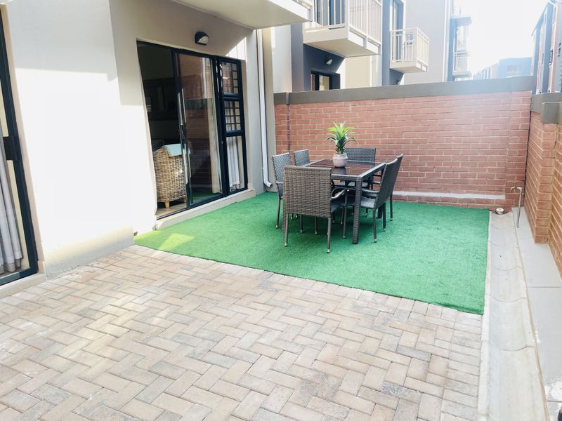 A private ground-floor patio featuring paved flooring, a section of artificial turf, and a brick boundary wall, accessible via sliding glass doors.