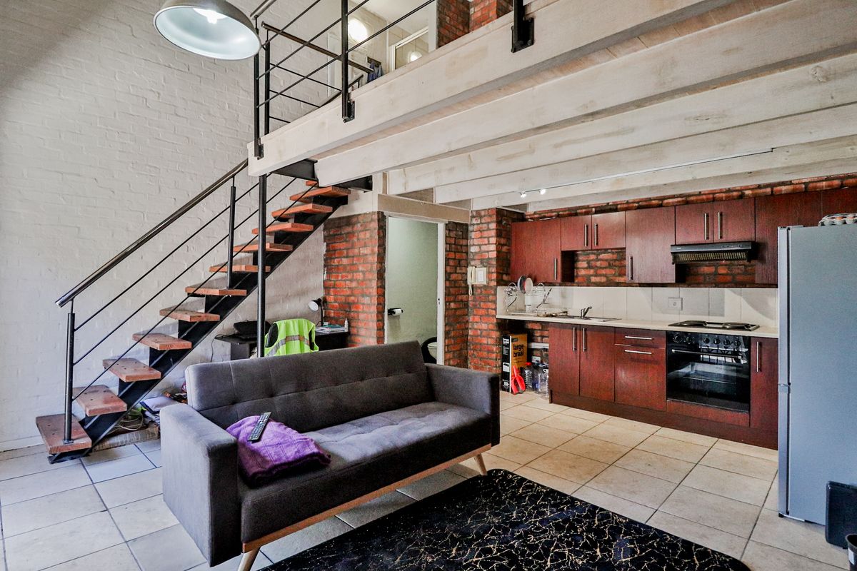Open-plan living area featuring a mezzanine level, exposed brick walls, a timber staircase, and a functional kitchen with built-in cabinetry and appliances.