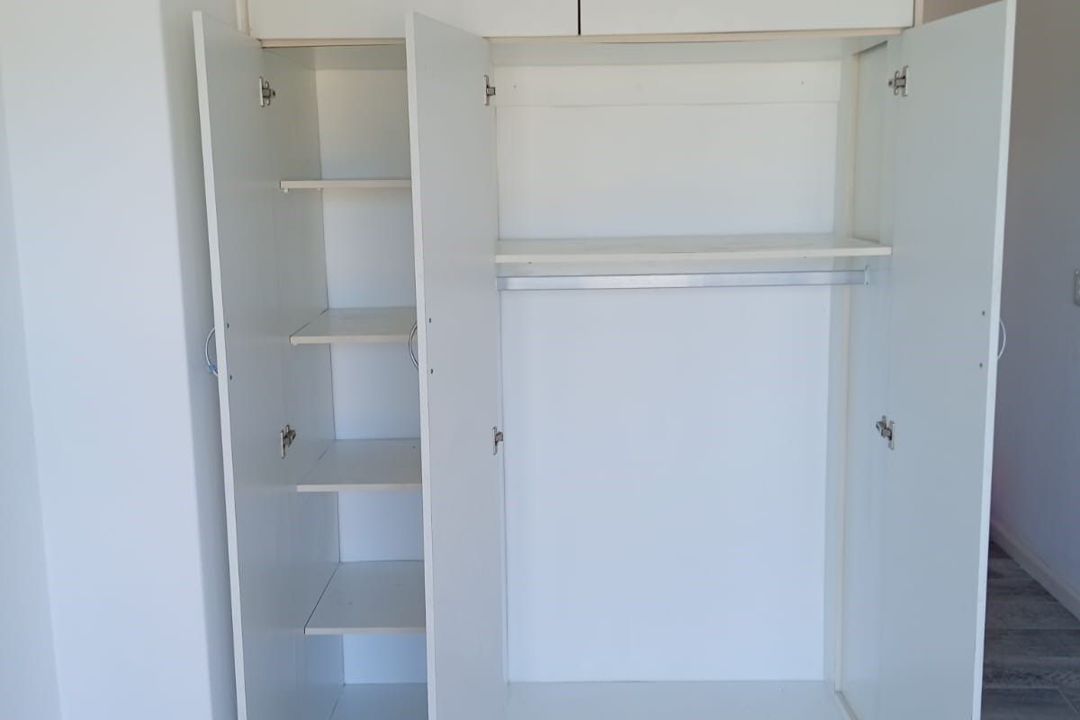 A clean, empty bedroom featuring a white built-in wardrobe with overhead storage, hanging space, and shelving, complemented by modern grey tiled flooring.