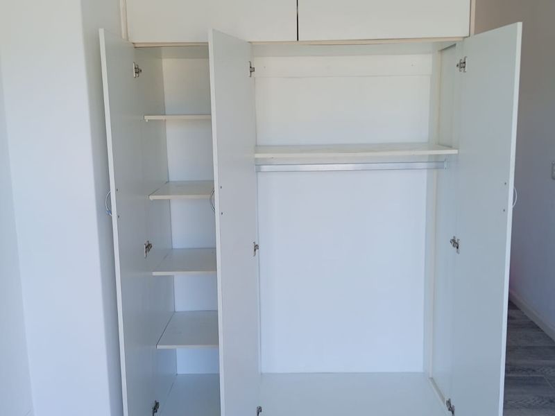 A clean, empty bedroom featuring a white built-in wardrobe with overhead storage, hanging space, and shelving, complemented by modern grey tiled flooring.