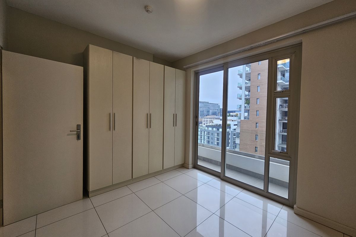 A bright bedroom featuring large format floor tiles, built-in floor-to-ceiling wardrobes, and a sliding glass door providing access to a balcony with city views.