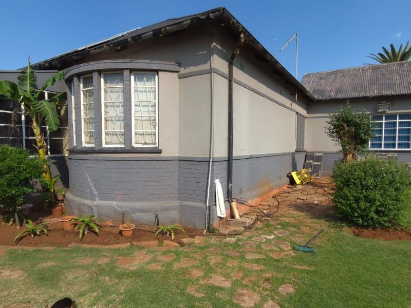 Rear view of a single-story house featuring a bay window, corrugated roof, and garden area with visible exterior maintenance required.