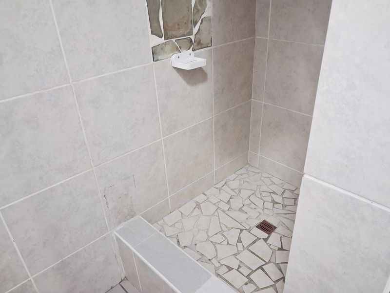 A shower enclosure featuring light-toned wall tiles and a mosaic-tiled floor, currently showing signs of wear and incomplete tiling in the niche area.