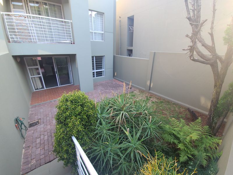 A rear view of the apartment featuring a paved patio area, a small garden, and an upper-level balcony with metal railings.