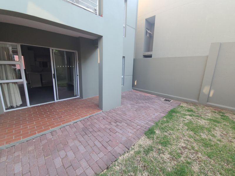 A private ground floor patio area featuring brick paving, a sliding door entrance, and a small adjacent garden space with a boundary wall.