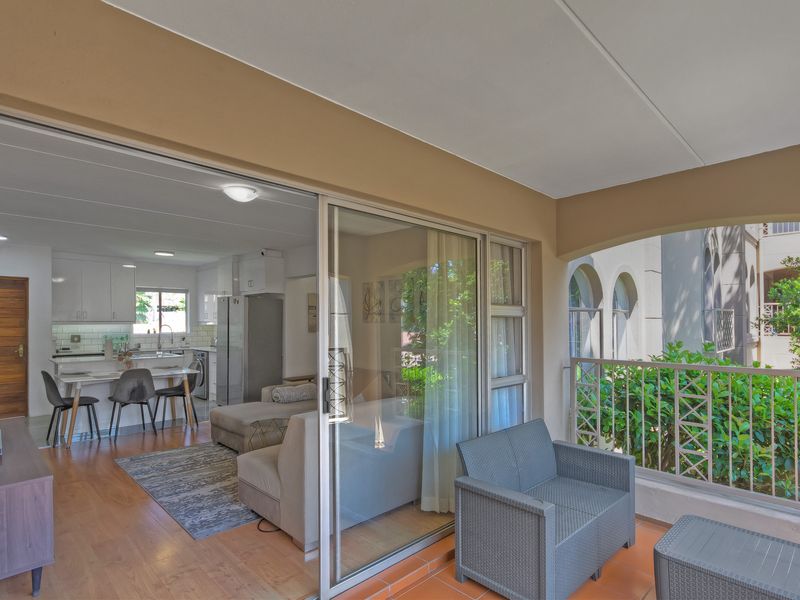 A covered balcony with tiled flooring and metal railing, providing seamless access to an open-plan living area and kitchen.