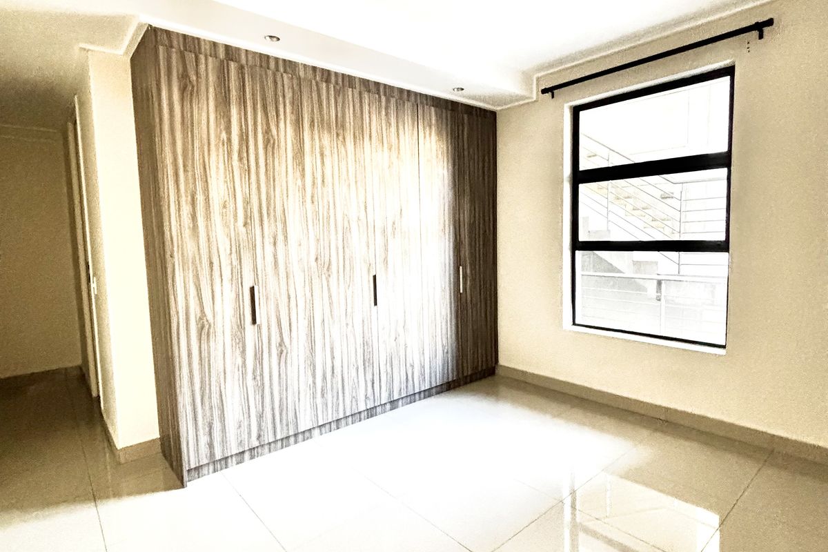 A bright, empty bedroom featuring large format floor tiles, a modern built-in wardrobe with wood-grain finish, and a window with black frame.