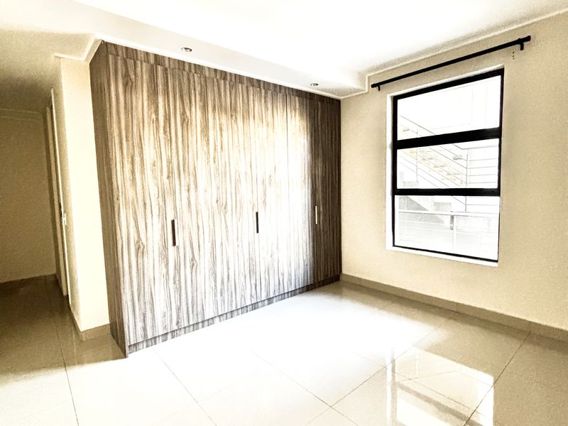 A bright, empty bedroom featuring large format floor tiles, a modern built-in wardrobe with wood-grain finish, and a window with black frame.