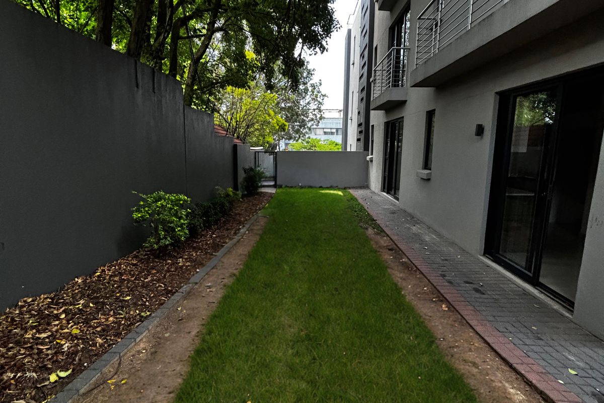 A narrow rear garden area featuring a grass lawn, paved walkway, and boundary wall, adjacent to the apartment building's exterior facade.