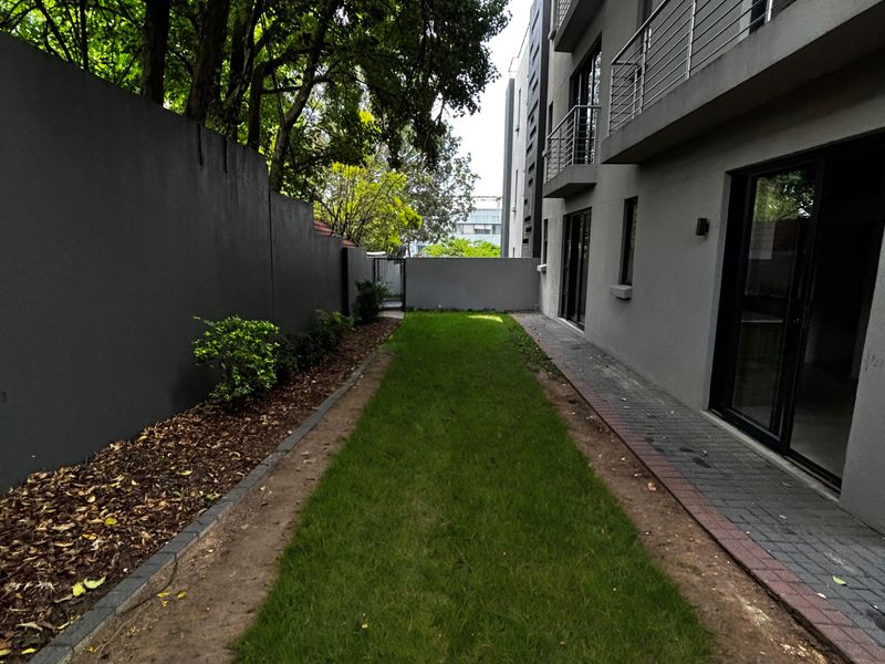 A narrow rear garden area featuring a grass lawn, paved walkway, and boundary wall, adjacent to the apartment building's exterior facade.