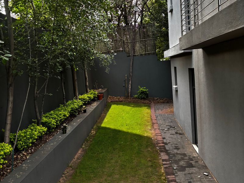 A private rear garden featuring a paved walkway, a small lawn area, and a raised planter bed with established trees and shrubs.