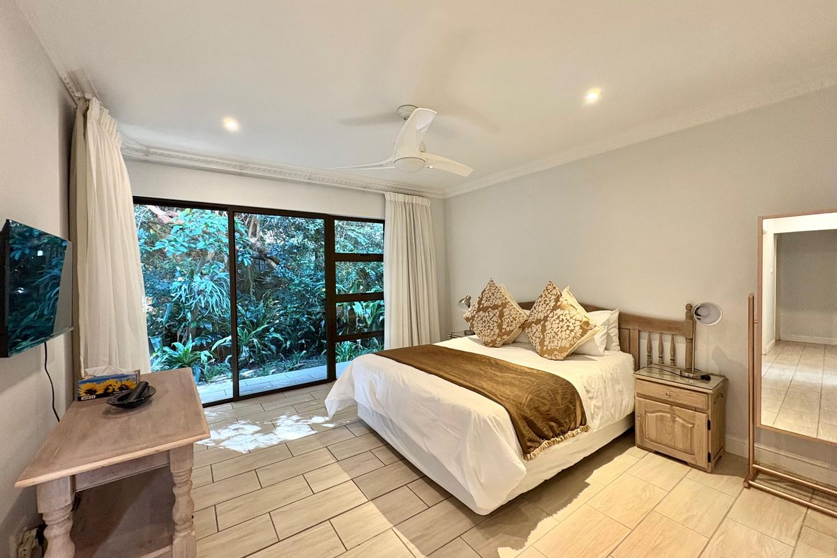 A bright bedroom featuring large sliding glass doors that open to a lush garden view, complemented by tiled flooring and a ceiling fan.