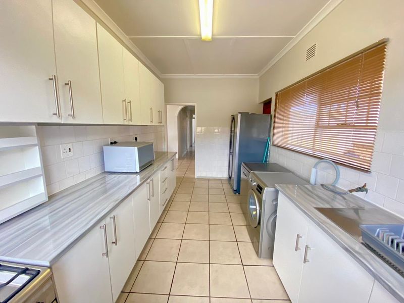 A functional galley-style kitchen featuring white cabinetry, laminate countertops, tiled flooring, and a tiled backsplash with integrated sink and appliance connections.