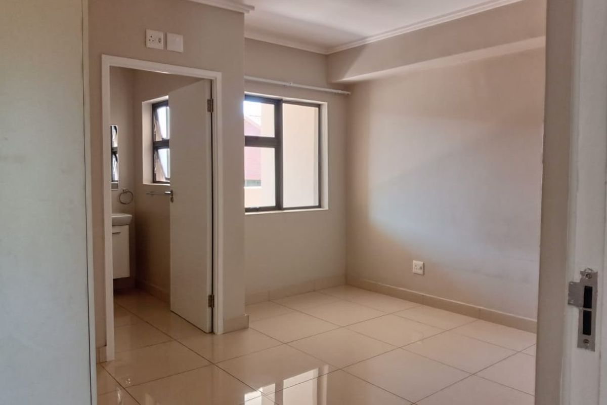 A bright, empty bedroom featuring neutral tiled flooring, clean walls, and an adjoining en-suite bathroom with a window.