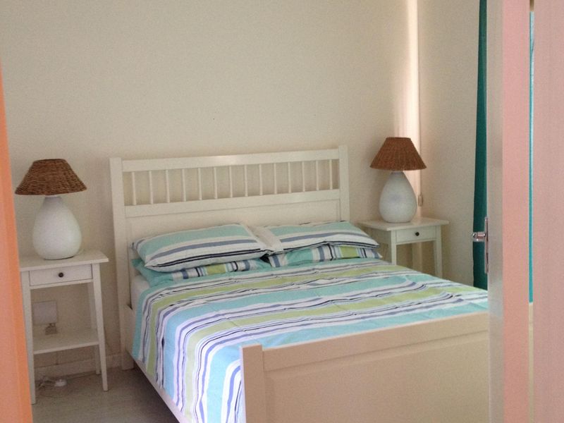 A simple, well-lit bedroom featuring light-toned laminate flooring and neutral wall finishes, viewed through a doorway.