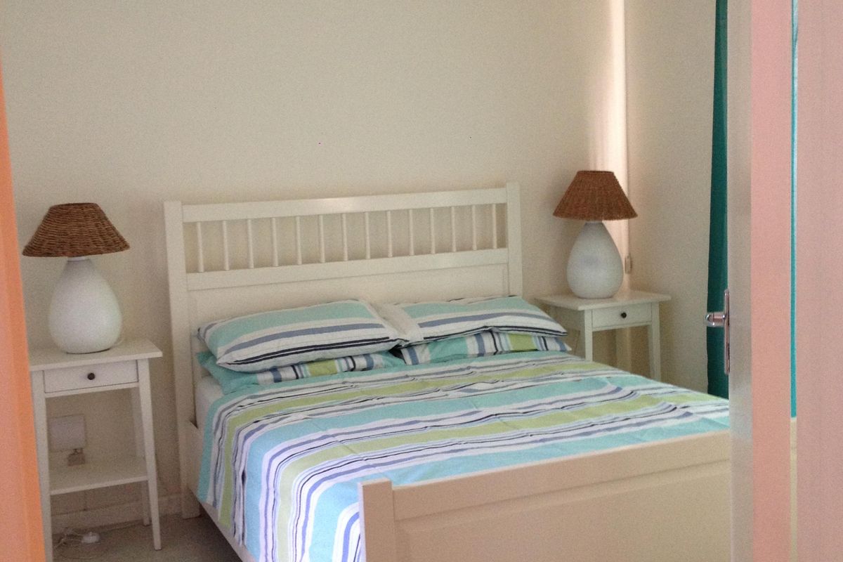 A simple, well-lit bedroom featuring light-toned laminate flooring and neutral wall finishes, viewed through a doorway.