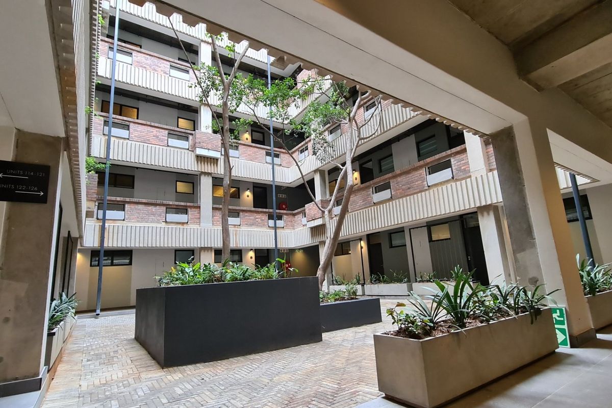A central open-air courtyard within an apartment complex, featuring brick paving, built-in concrete planters, and multi-level walkways with exposed concrete and brick finishes.