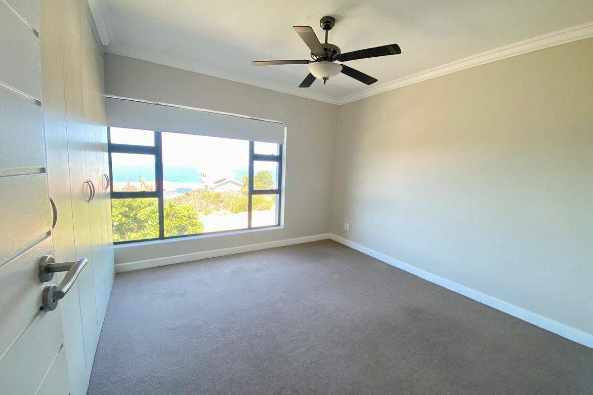 A bright, empty bedroom featuring neutral carpeting, built-in cupboards, a ceiling fan, and a large window with views of the surrounding area.