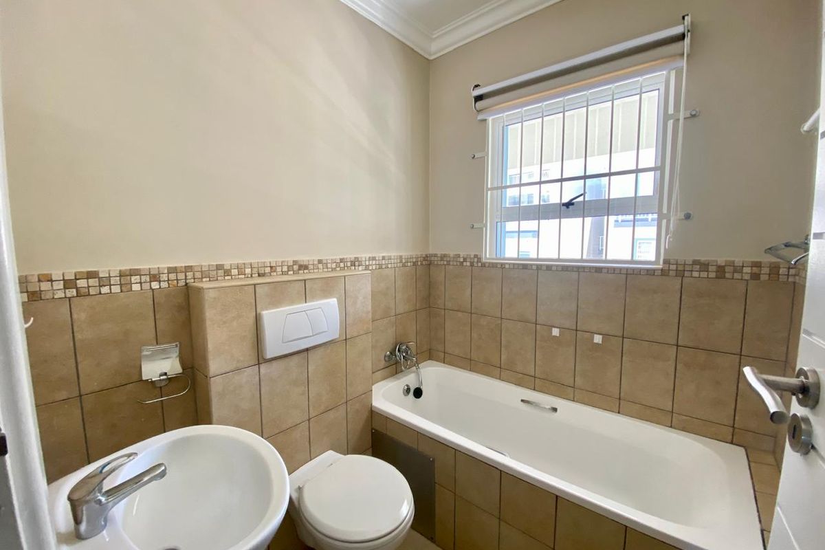A functional bathroom featuring a built-in bathtub, wall-mounted toilet, and pedestal basin with neutral wall tiling and security bars on the window.
