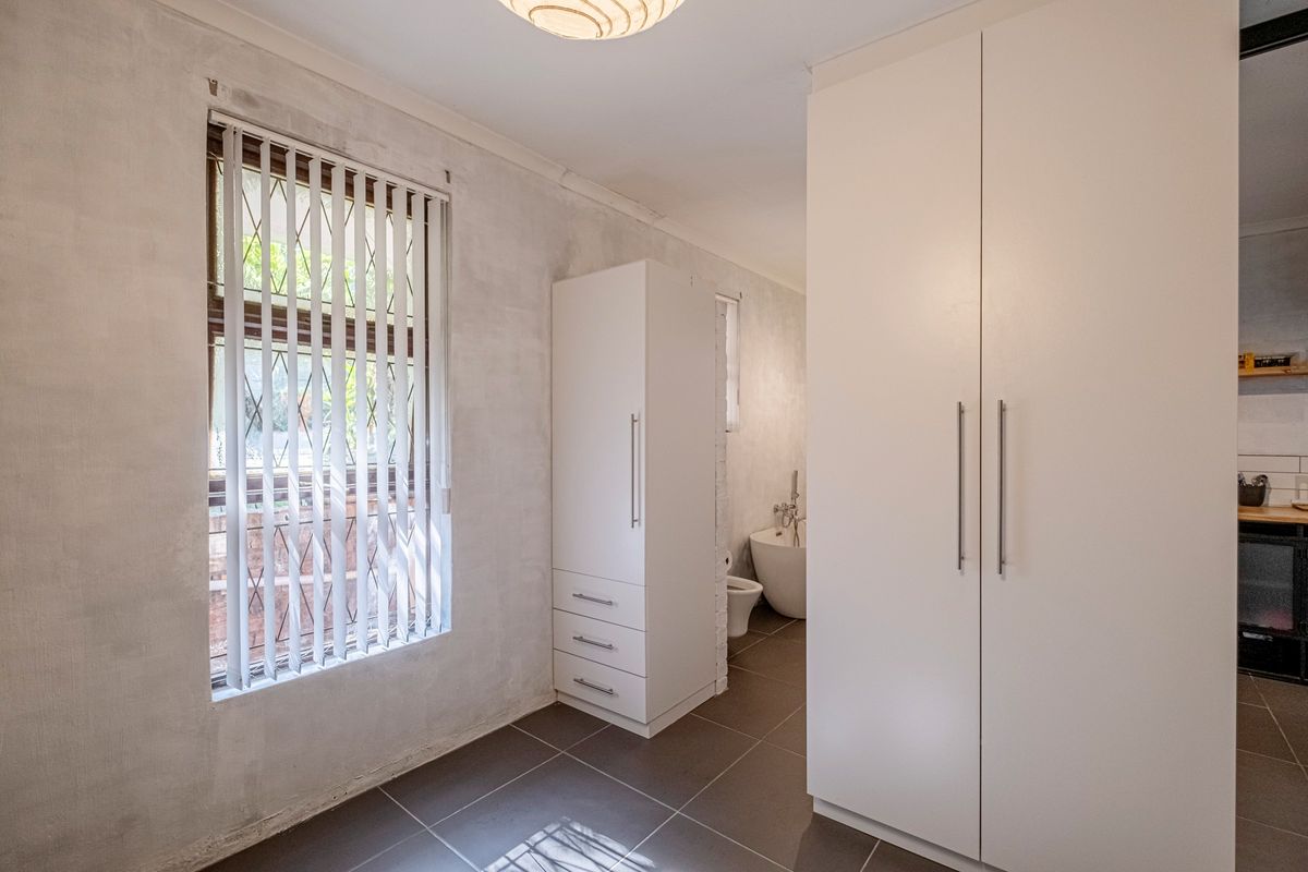 A bedroom featuring tiled flooring, built-in cupboards, and vertical blinds, with an adjacent view into a bathroom area.