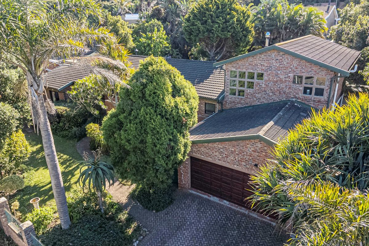 An elevated aerial view of a multi-level brick apartment building featuring a tiled roof, paved driveway, double garage, and established garden surroundings.