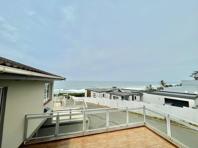 An elevated balcony featuring a modern glass balustrade and tiled flooring, offering expansive views of the surrounding estate and the ocean.