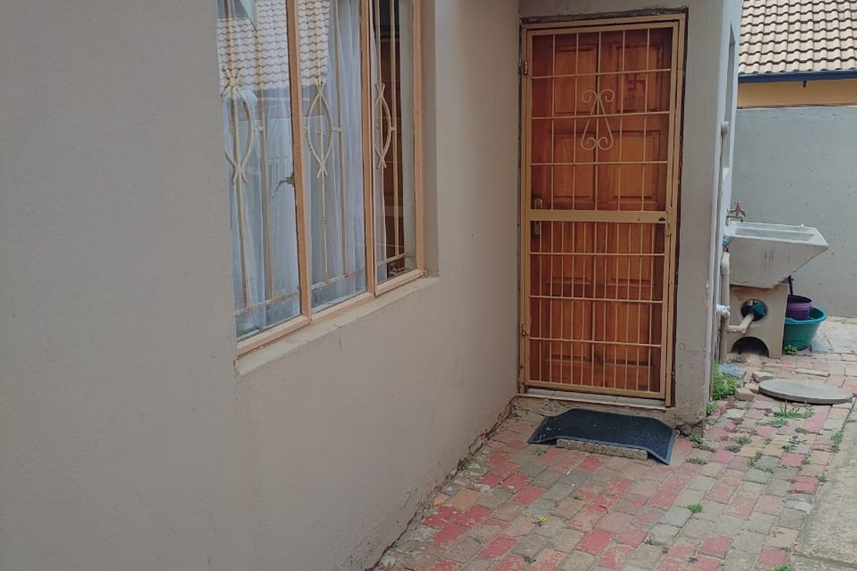 Exterior rear view showing a paved courtyard area with a utility sink, security gate, and window with burglar bars.