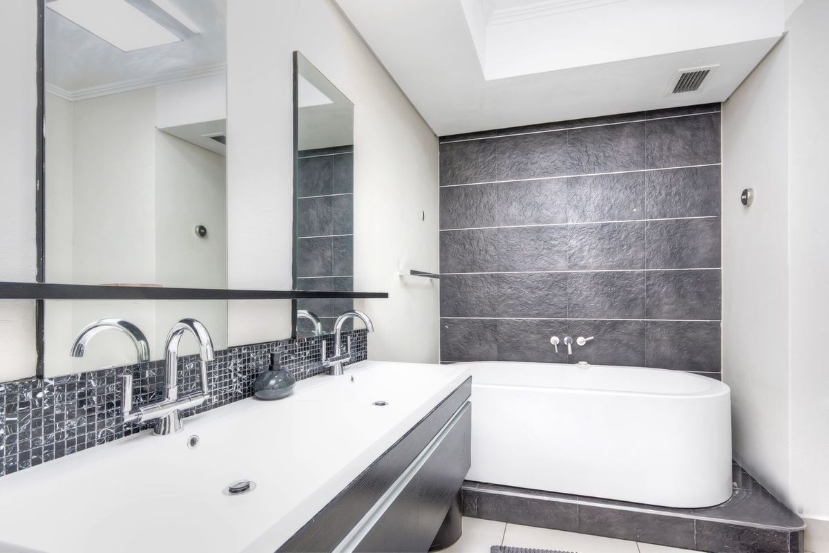Modern bathroom featuring a double vanity with chrome fixtures, a deep soaking bathtub, and contrasting dark tiled feature wall.