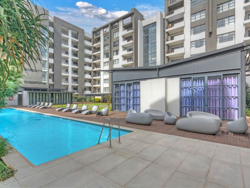 A well-maintained communal swimming pool area featuring a wooden deck, paved surround, and modern apartment building architecture in the background.