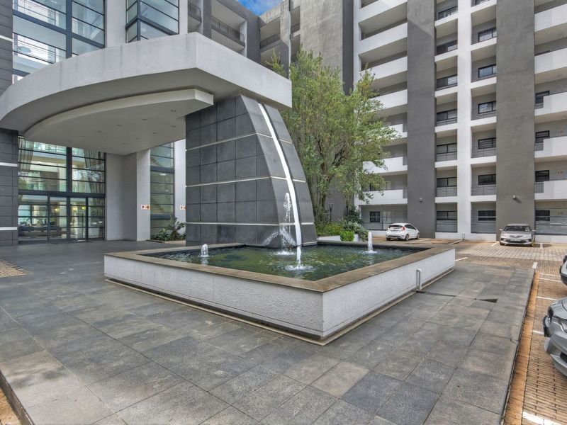 Modern apartment complex entrance featuring a prominent architectural water feature, paved driveway, and covered porte-cochère.
